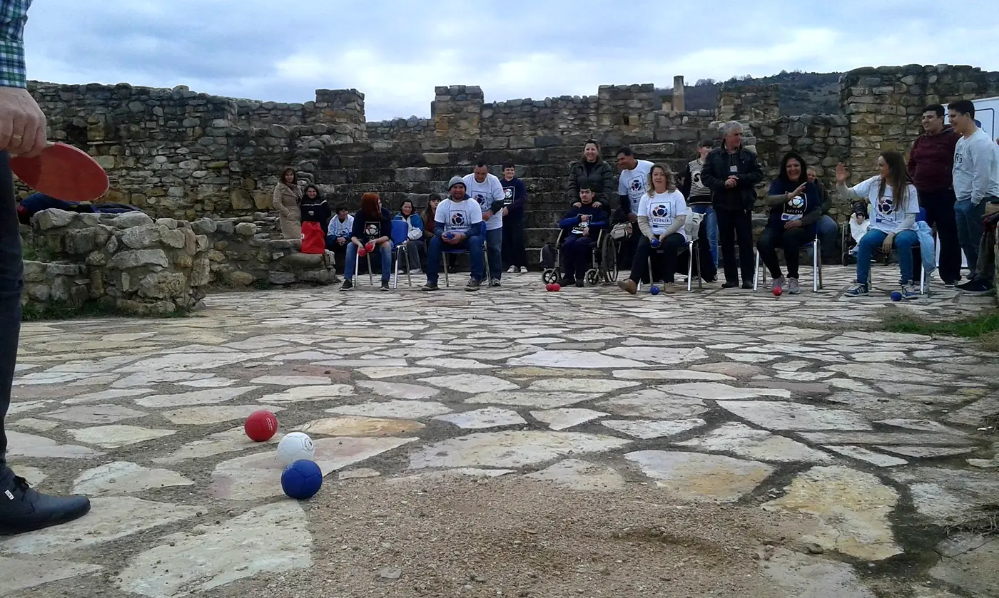 A Boccia match in play: red, white and blue balls on a stone court, players seated around the edge, two of them using wheelchairs.