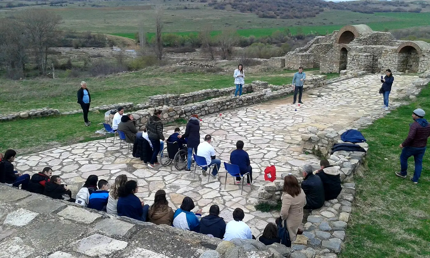 A Boccia session under way on a stone court at an open-air archaeological site, with players and spectators seated around the court and a green valley behind